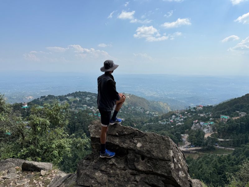 Man standing on a rocky ledge overlooking McLeod Ganj and the Kangra Valley, Himachal Pradesh Man standing on a rocky ledge overlooking McLeod Ganj and the Kangra Valley, Himachal Pradesh, during a scenic hike near Dharamkot on a clear day.