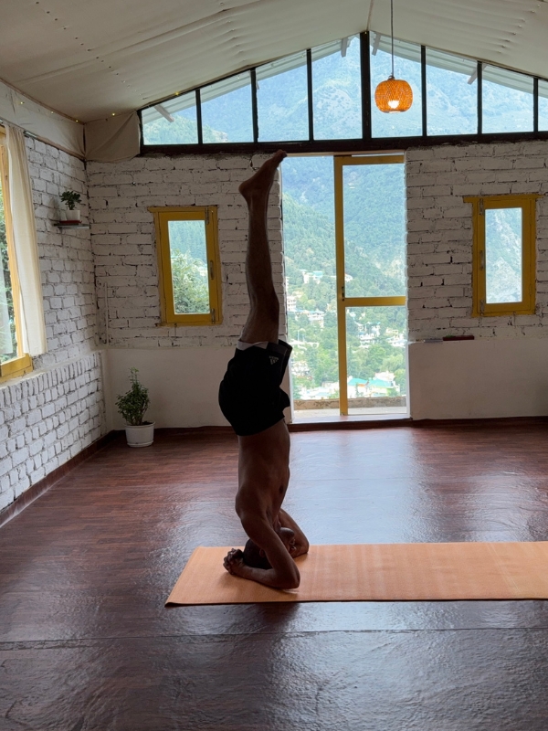 Man practicing headstand yoga pose (Sirsasana) on a mat inside a mountain-view studio in Dharamkot, surrounded by white brick walls and large windows overlooking the Dhauladhar range. 